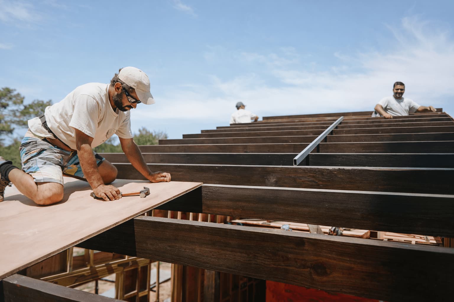 Carpenter laying panels on the roof beams of a timber house, two colleagues working behind him.