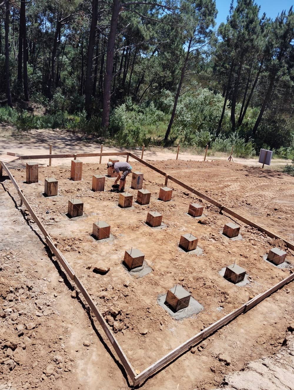 Foundation grid of concrete piers with rebar anchors and string lines, a worker checking the alignment.