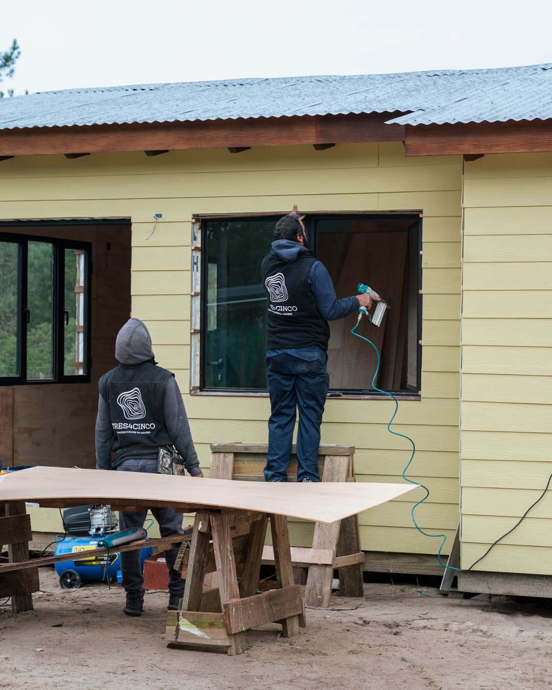 Two workers in TRES4CINCO jackets working on a window of the yellow facade from outside, a panel on trestles in front.