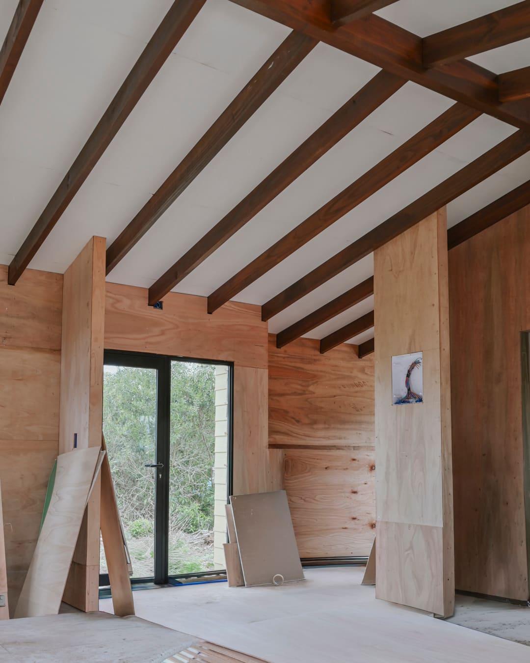 Room lined with timber panels, dark roof beams, glass door to the outside, panels leaning against the wall.