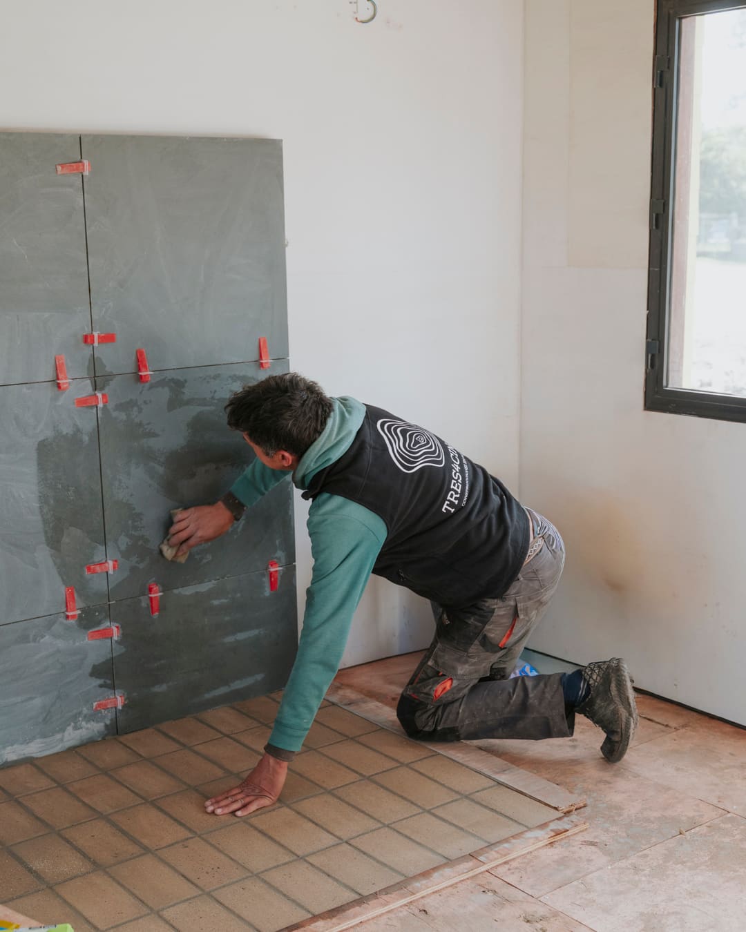 Worker setting large grey wall tiles with levelling clips, small tiles on the floor.