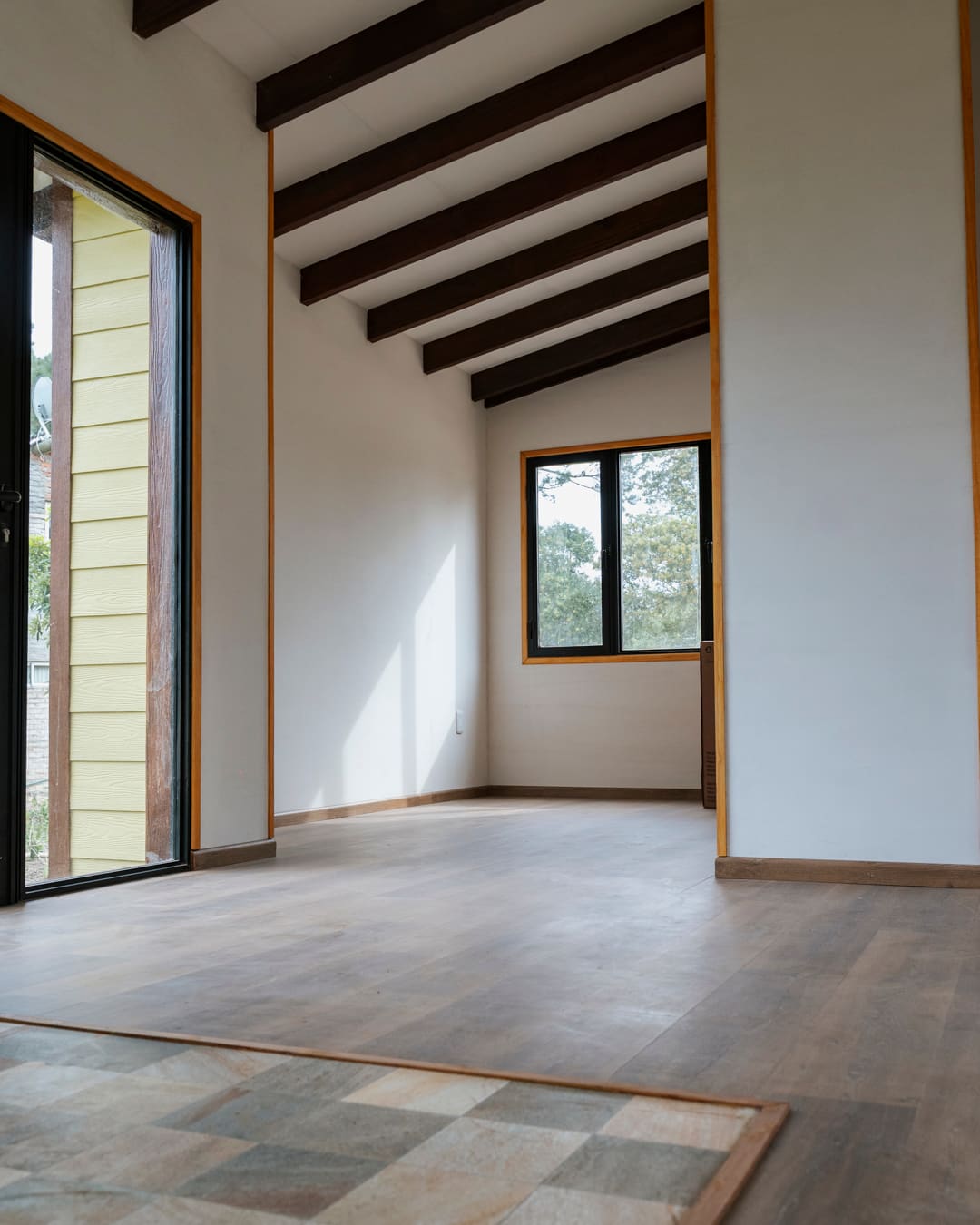 Living room with a timber floor and a tiled field, tall window to the deck and dark roof beams.