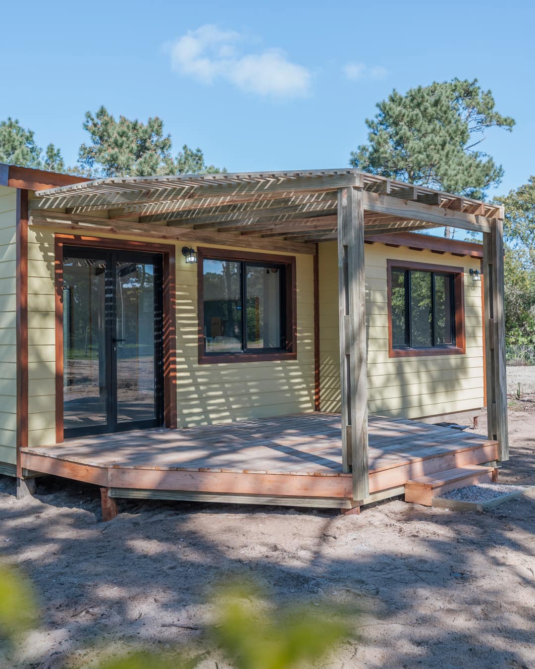 Corner of the house with a pergola over the deck, glass door and windows in the yellow facade, pine shade on the timber.