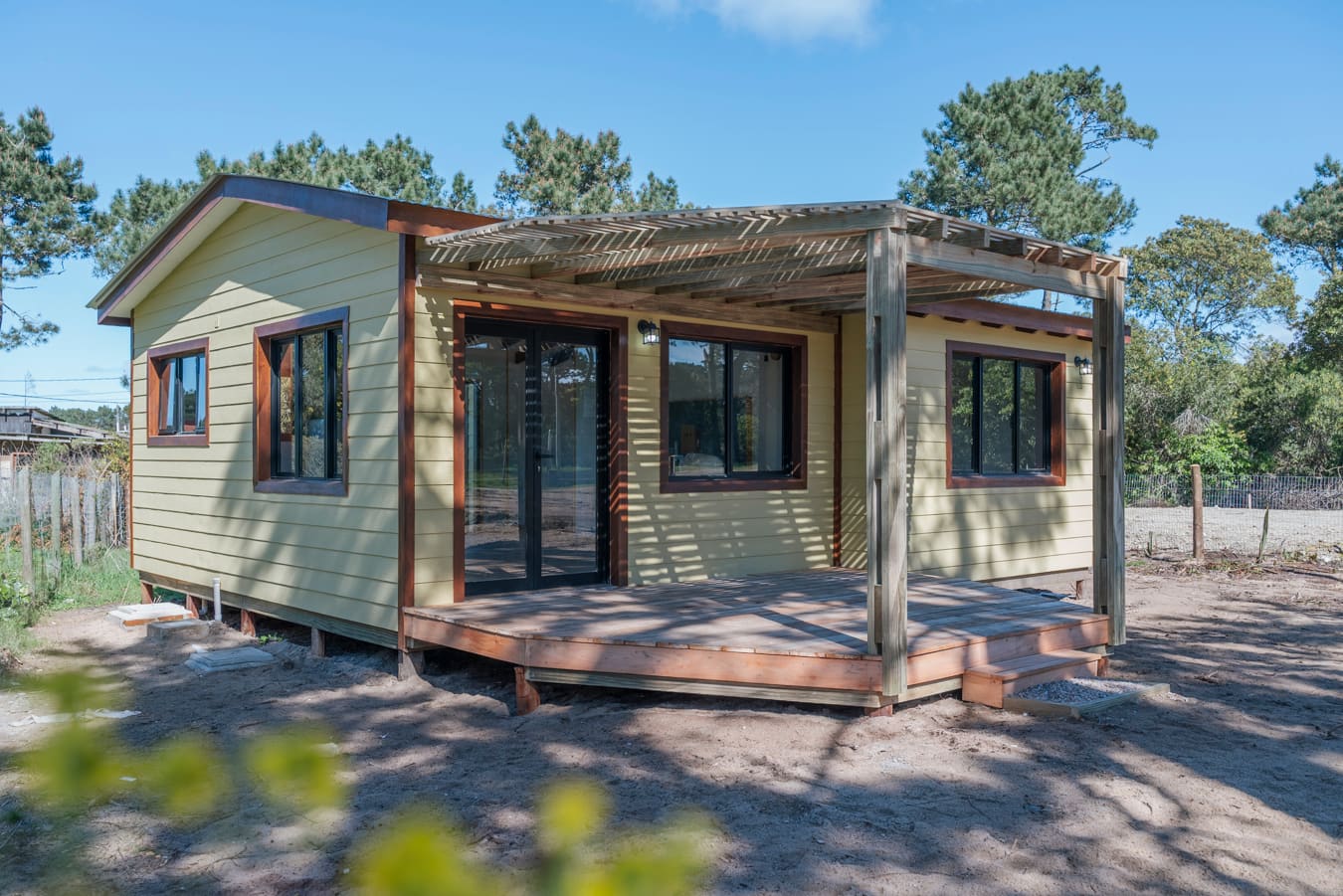 ILKA from outside: yellow timber house with a pergola over the deck, pines in the background.