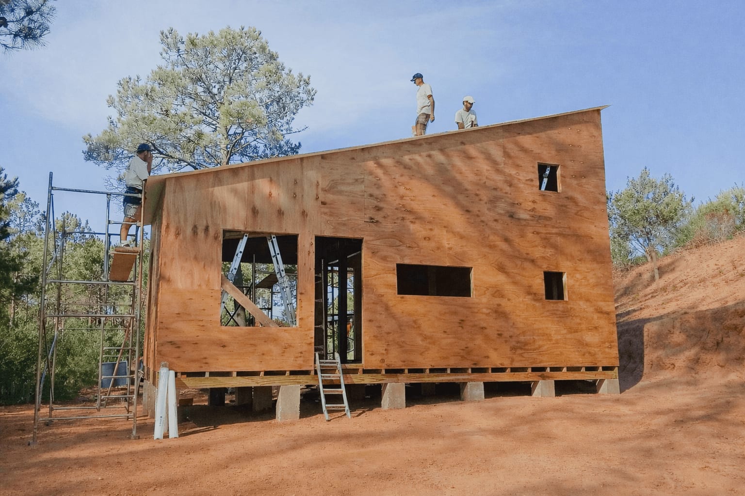 The team raises a timber frame wall on concrete piers, two workers on the roof.