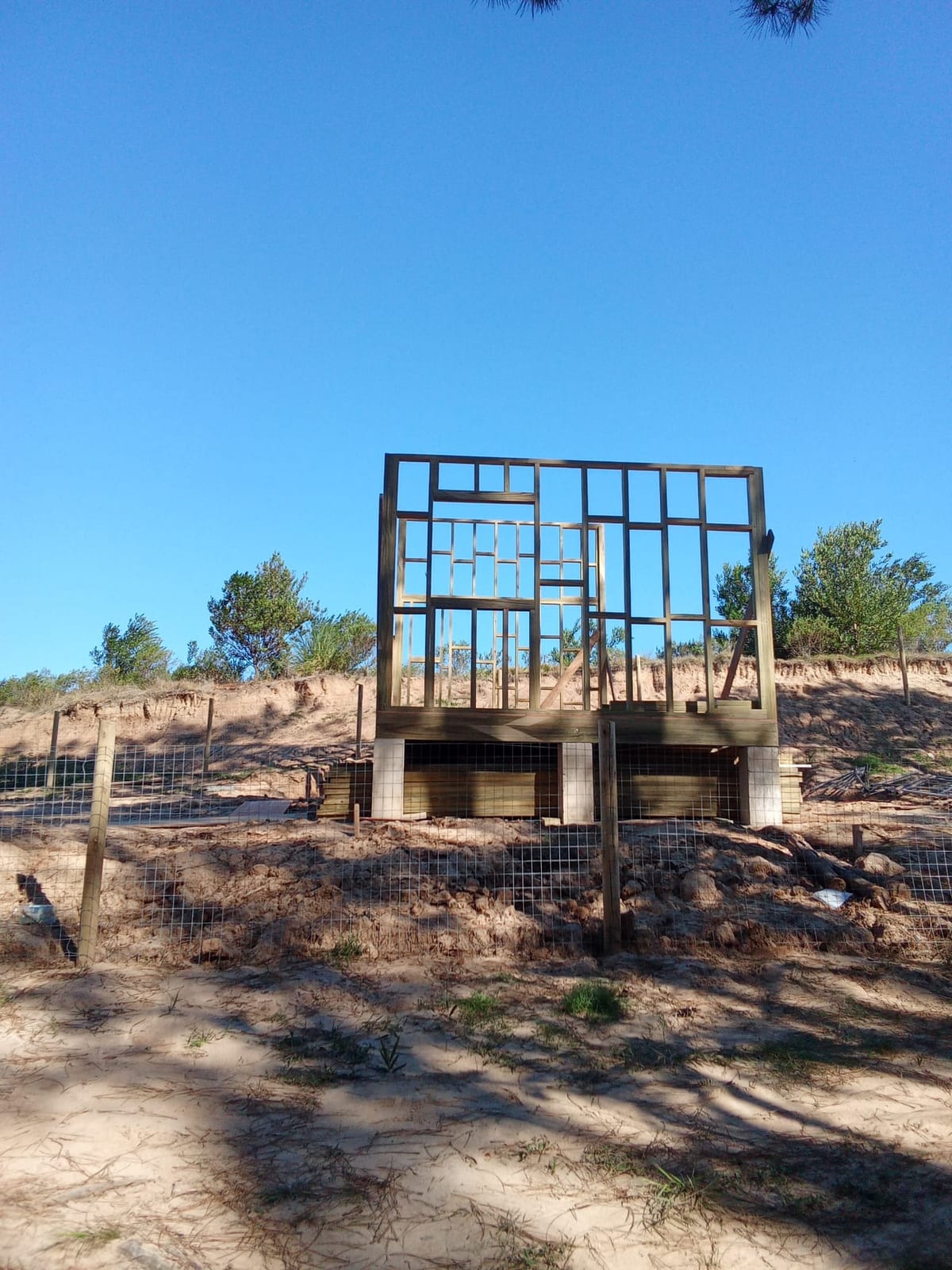 Freestanding timber frame skeleton against a deep blue sky on sandy coastal dunes.