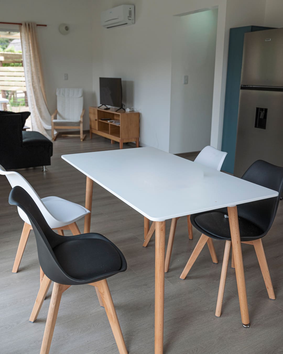 White dining table with four chairs, a TV unit, an armchair, a sofa and an air conditioner behind.
