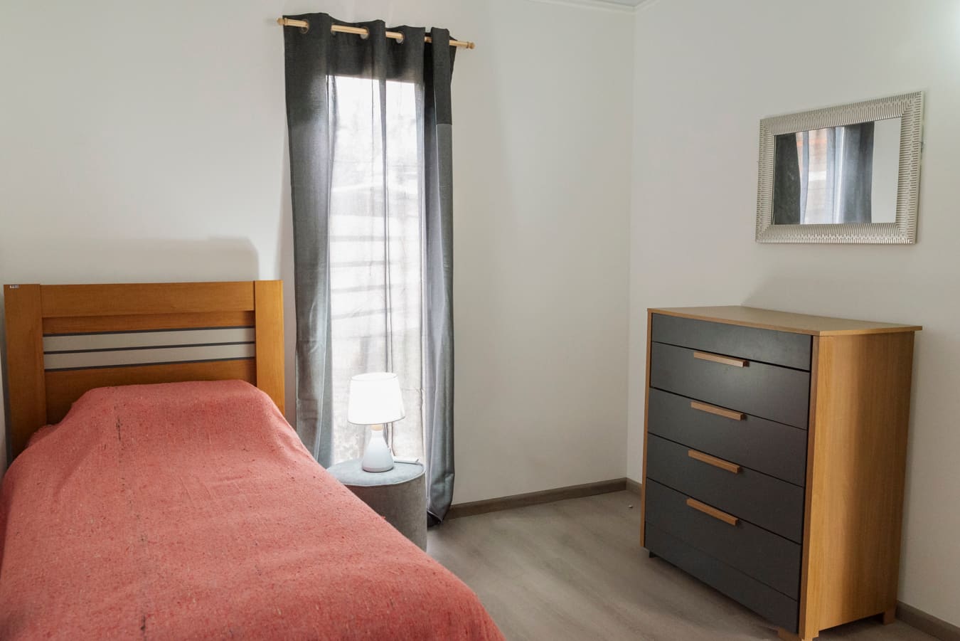 Single bed with a coral blanket beside a window with a grey curtain, a wooden chest of drawers and mirror at right.