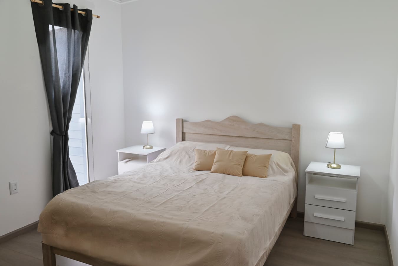 Double bed with beige cushions between two white bedside tables with lamps, a grey curtain at left.