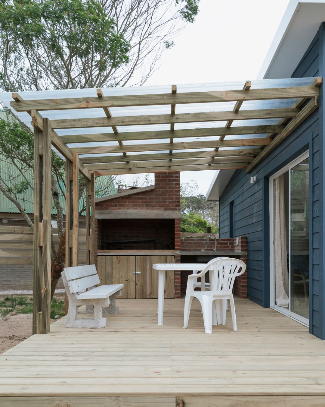 Pergola with a translucent roof over the timber deck, a bench, table and chair beneath, the brick barbecue behind.