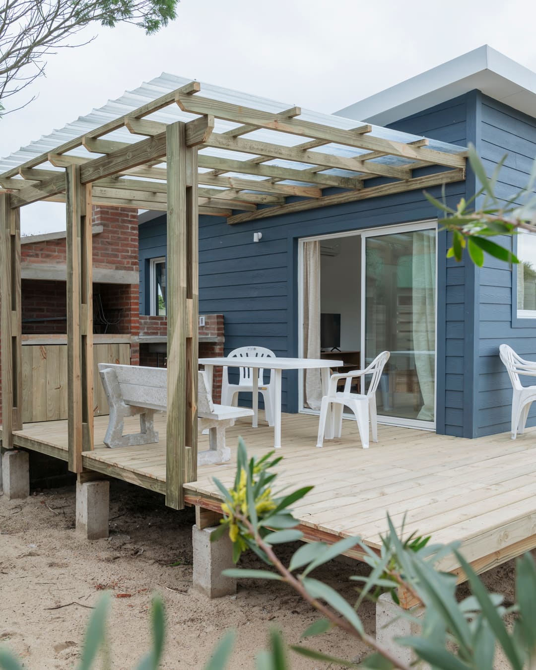 Timber deck under a pergola with a bench, a table and white chairs in front of the house's sliding glass door.