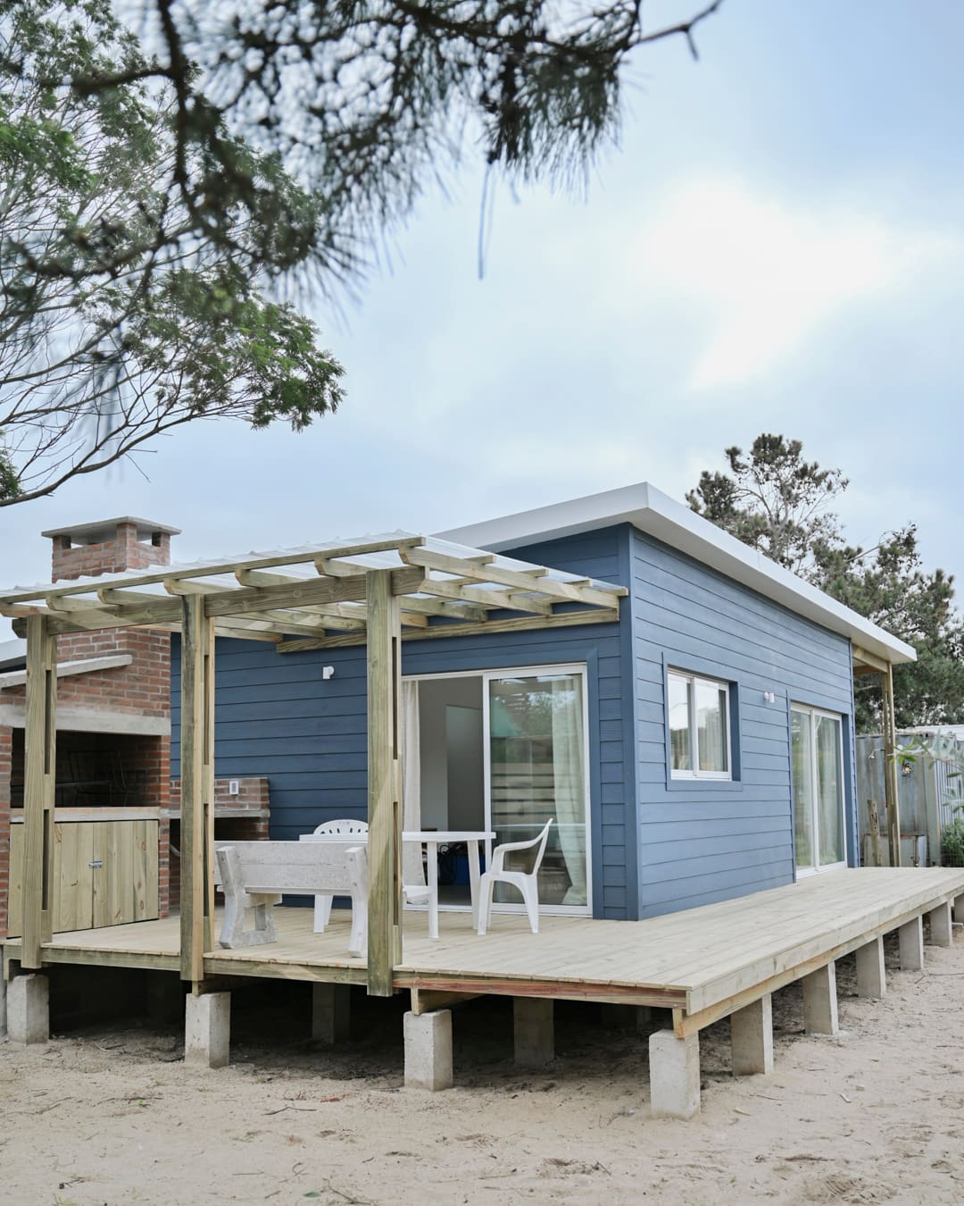 Blue wooden house on concrete footings with a deck and pergola, a brick barbecue with a chimney at left.