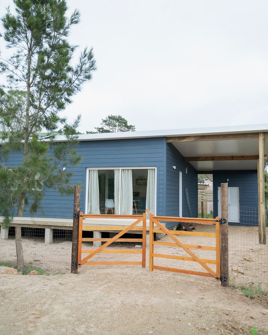 Double wooden gate in front of the blue house, with a covered passage at right and a pine tree at left.