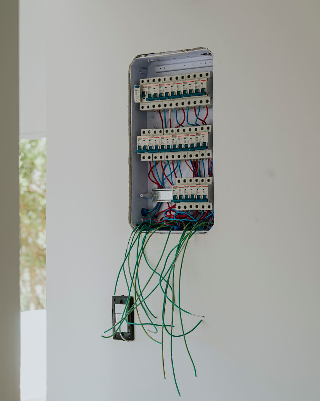Open electrical panel on a white wall with circuit breakers and many green wires hanging down.