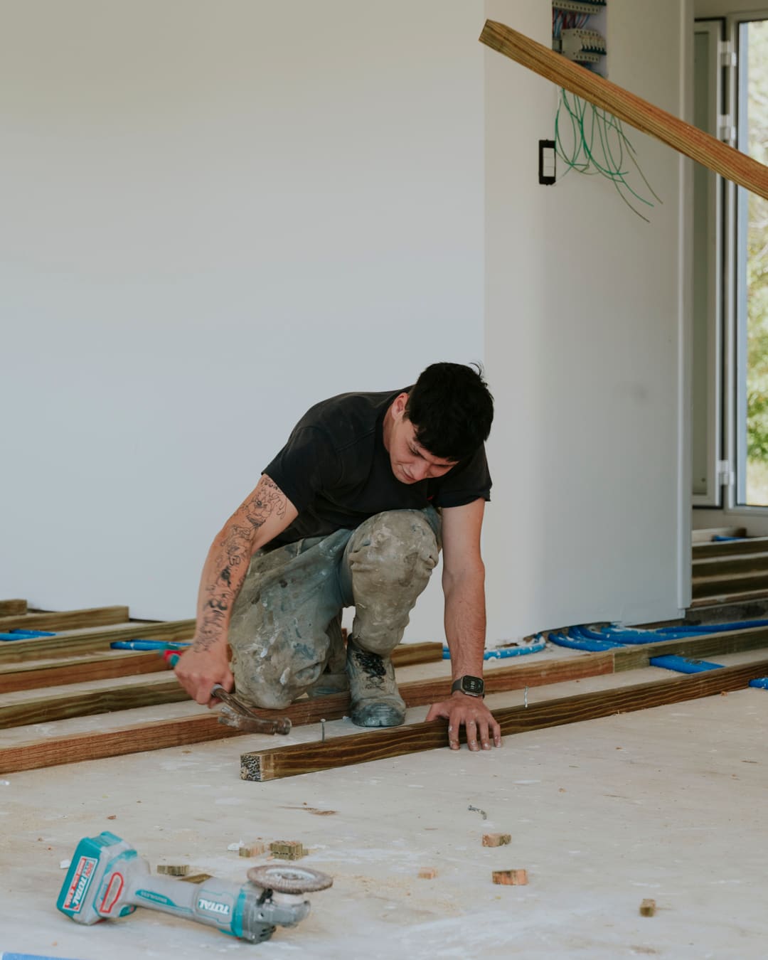 A worker kneels on the floor fixing wooden battens, with an angle grinder lying beside him.