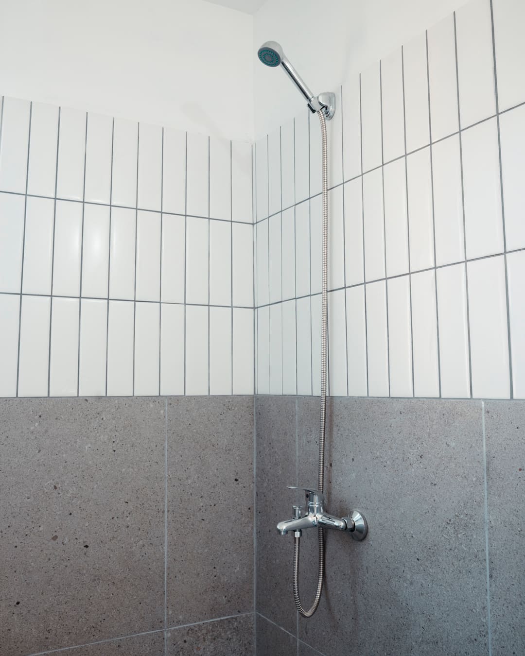 Shower with a handheld head, white vertical tiles above and grey stone-look tiles below.