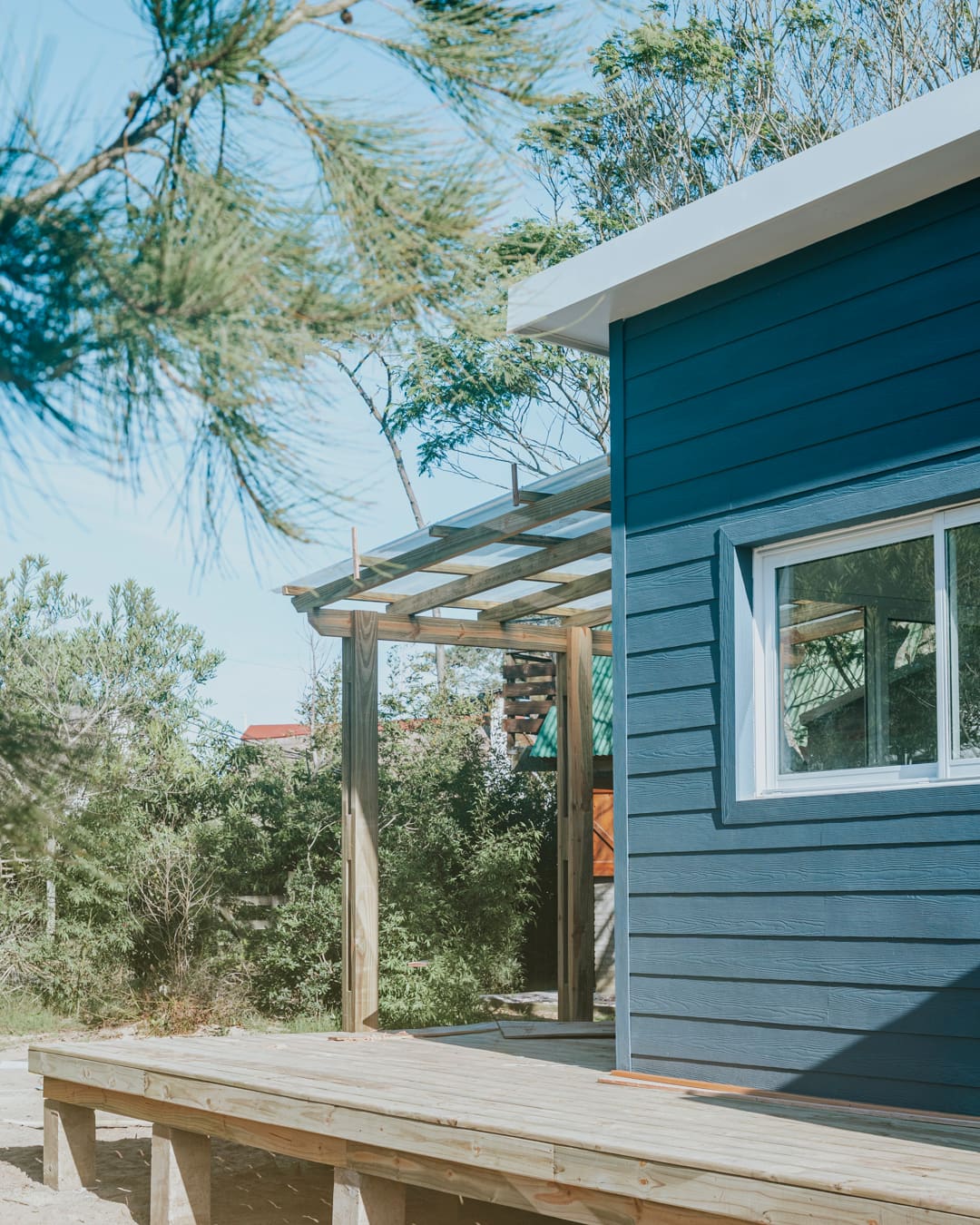 Blue wooden facade with a white window, a pergola and a timber deck beneath pine branches.