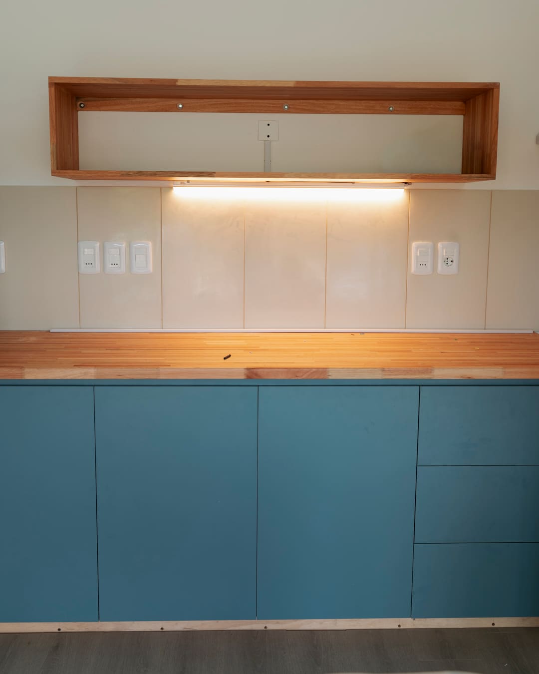 Teal base cabinets with a wooden worktop and a wooden shelf above lit by a warm light strip.