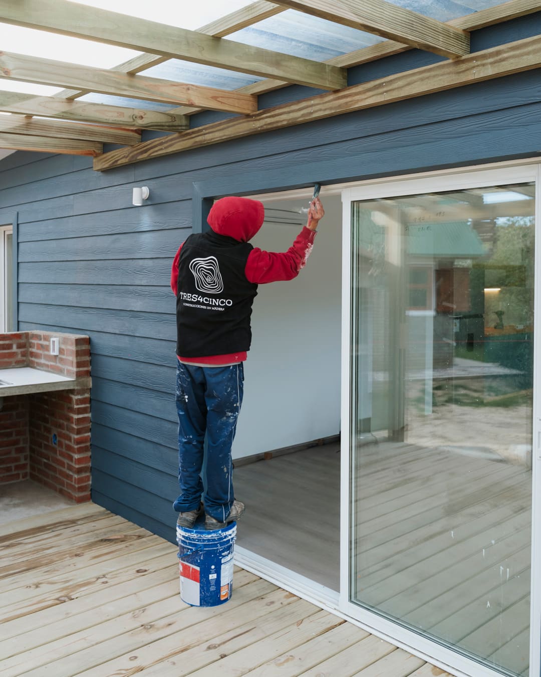 A worker standing on a paint bucket paints the blue facade next to the sliding glass door.