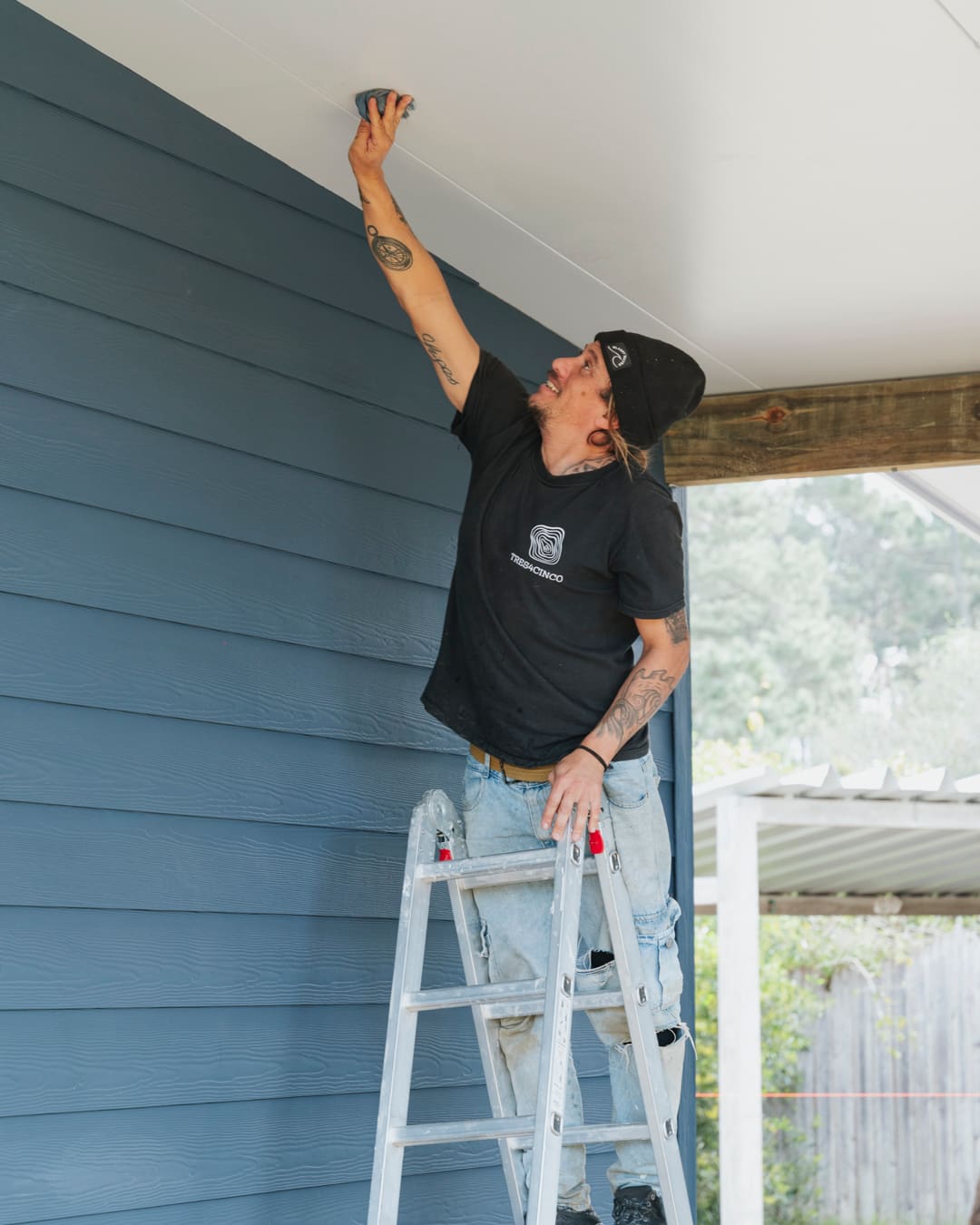 A worker on a ladder works on the white underside of the roof overhang beside the blue facade.