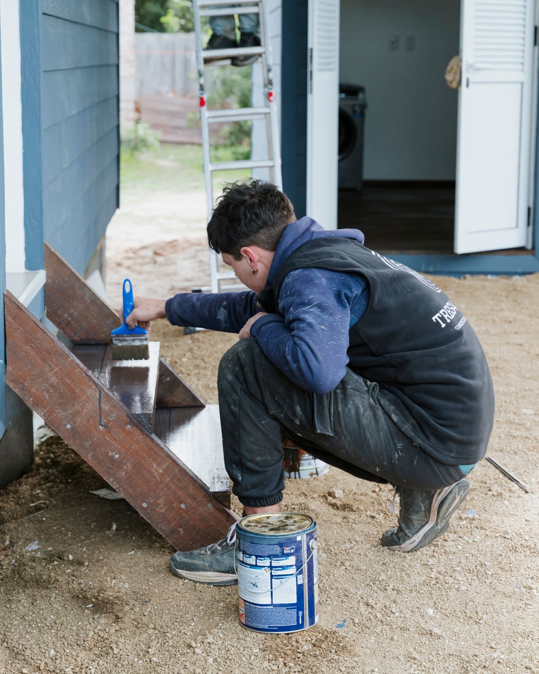 A worker crouches by a small wooden staircase and paints the steps, an open white door behind him.