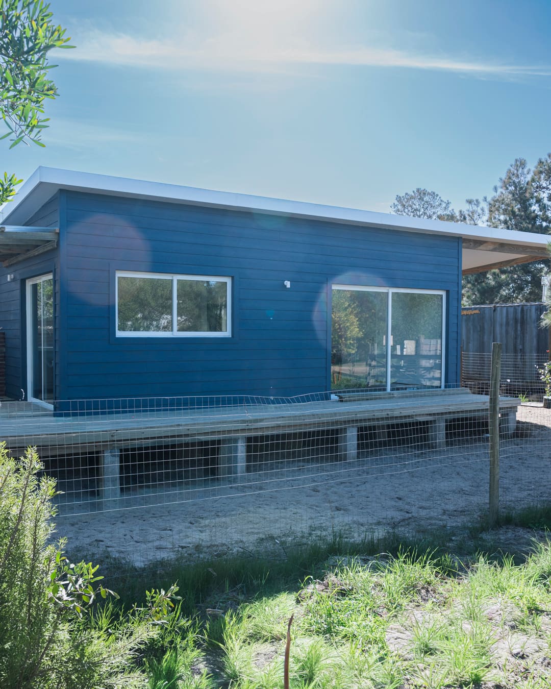 Blue wooden house with a deck behind a wire fence under a sunny sky with lens flare.