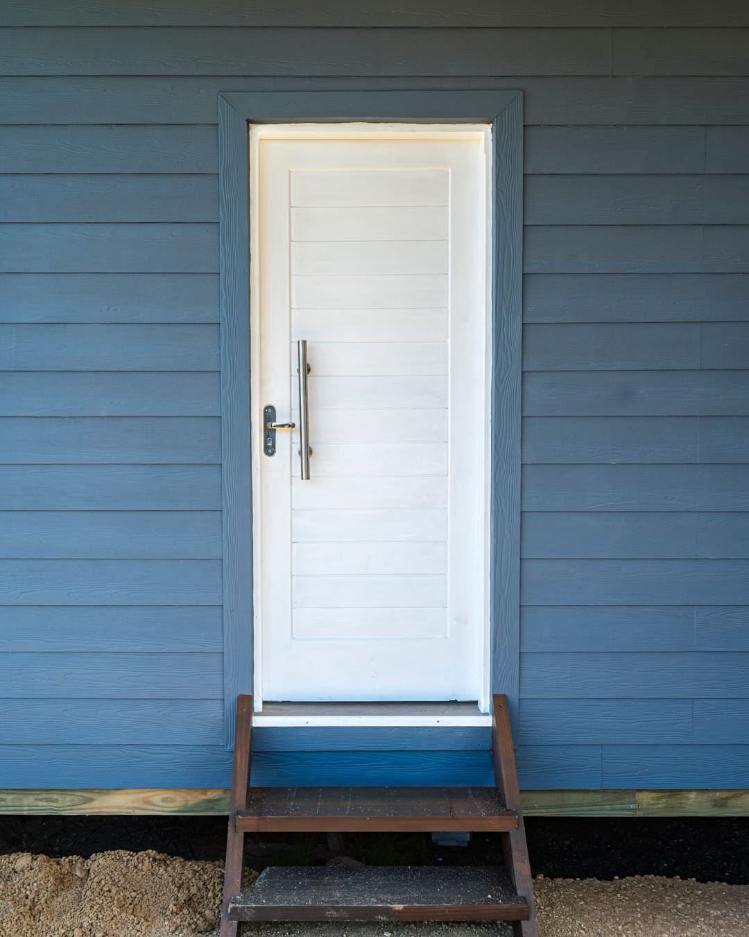 White door with a vertical handle in the blue-grey wooden facade, a small dark wooden stair in front.