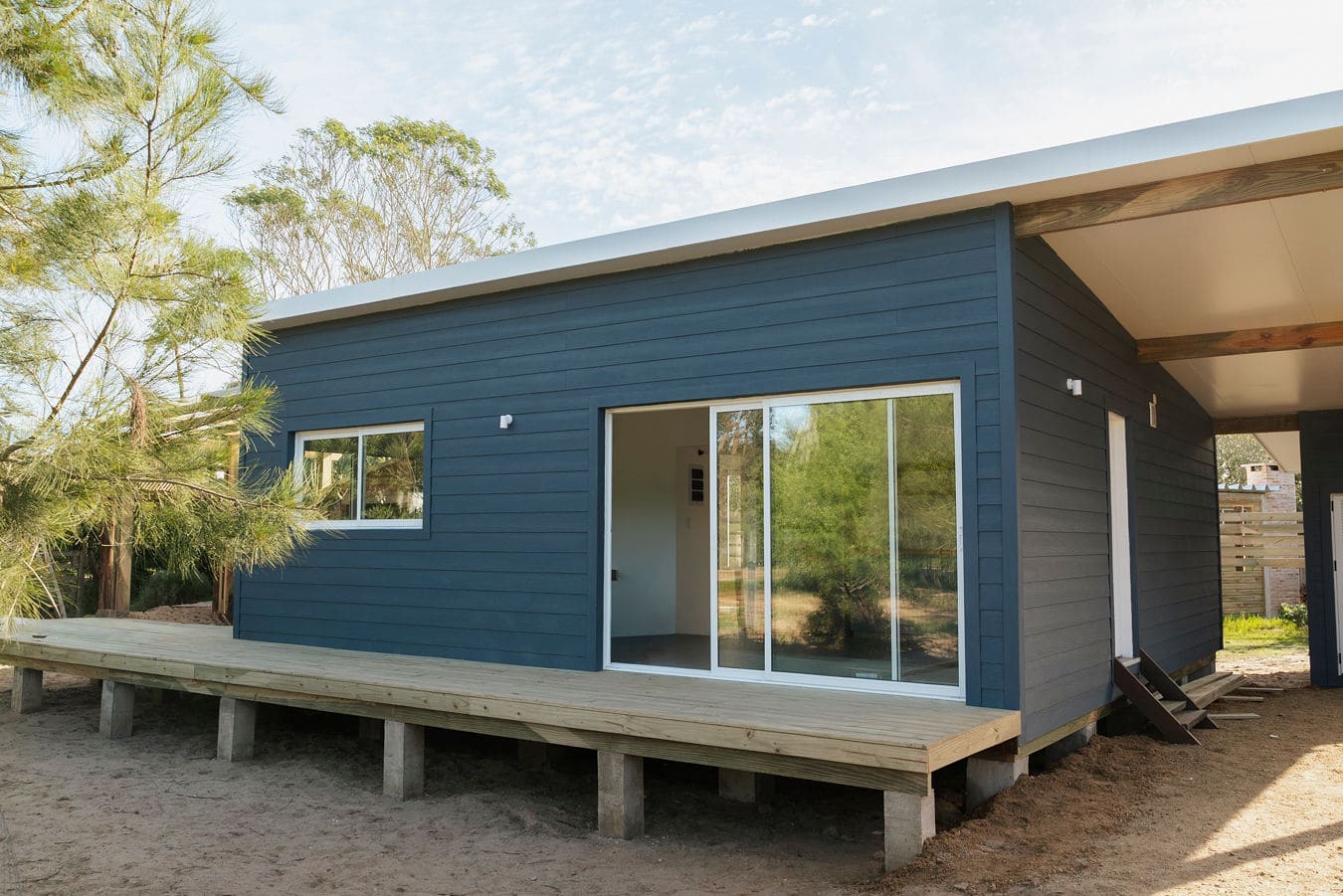Dark blue wooden house with a sliding glass door, a raised deck and a covered passage on the right.